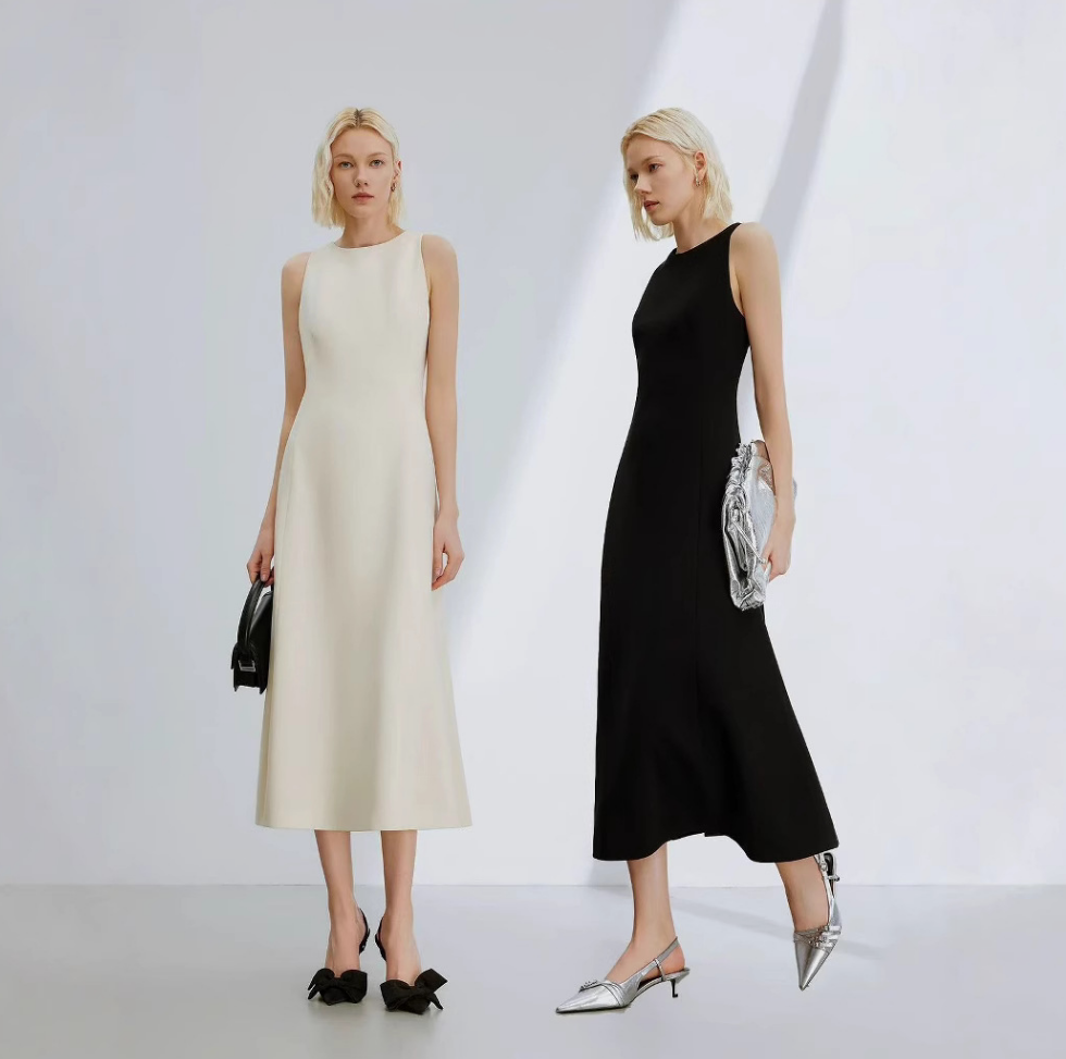 Two models standing in a studio wearing sleeveless midi dresses, one in cream and one in black, each holding a metallic clutch and wearing pointed heels.
