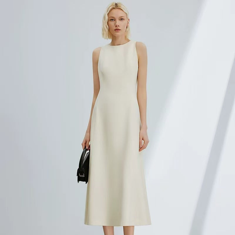 A woman modeling a sleeveless, high-neck midi dress in a warm off-white or cream color, holding a small black handbag against a minimalist light gray background with soft diagonal shadows.
